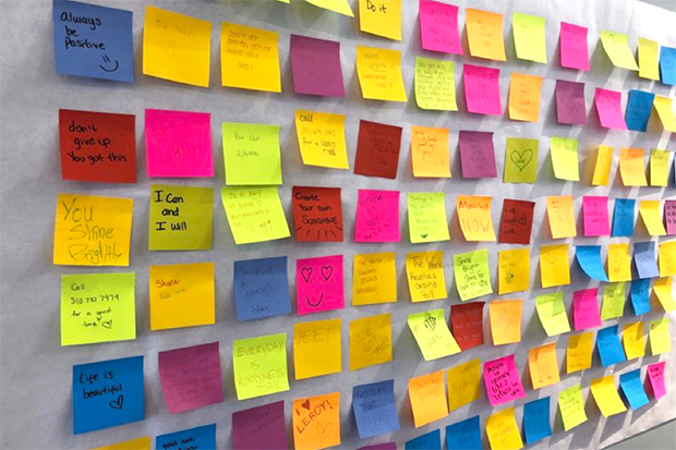 A wall with a grid of colorful post-it notes with drawings including  hearts, smiley faces and sunshine and kind phrases including: "I can and I will," "You shine brightly," "Always be positive." "Don't give up, you got this." "Life is beautiful."